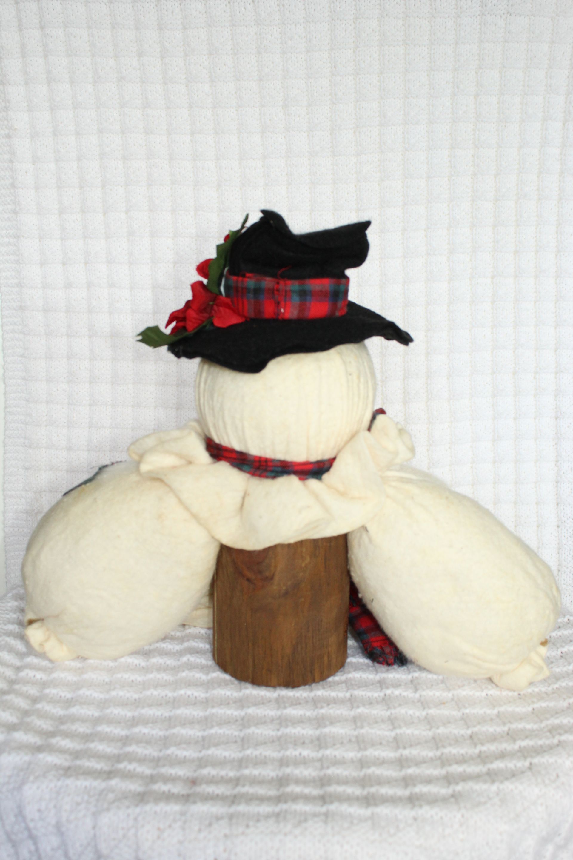 Snowman plush with black hat, red plaid scarf, and flower accent on a white background