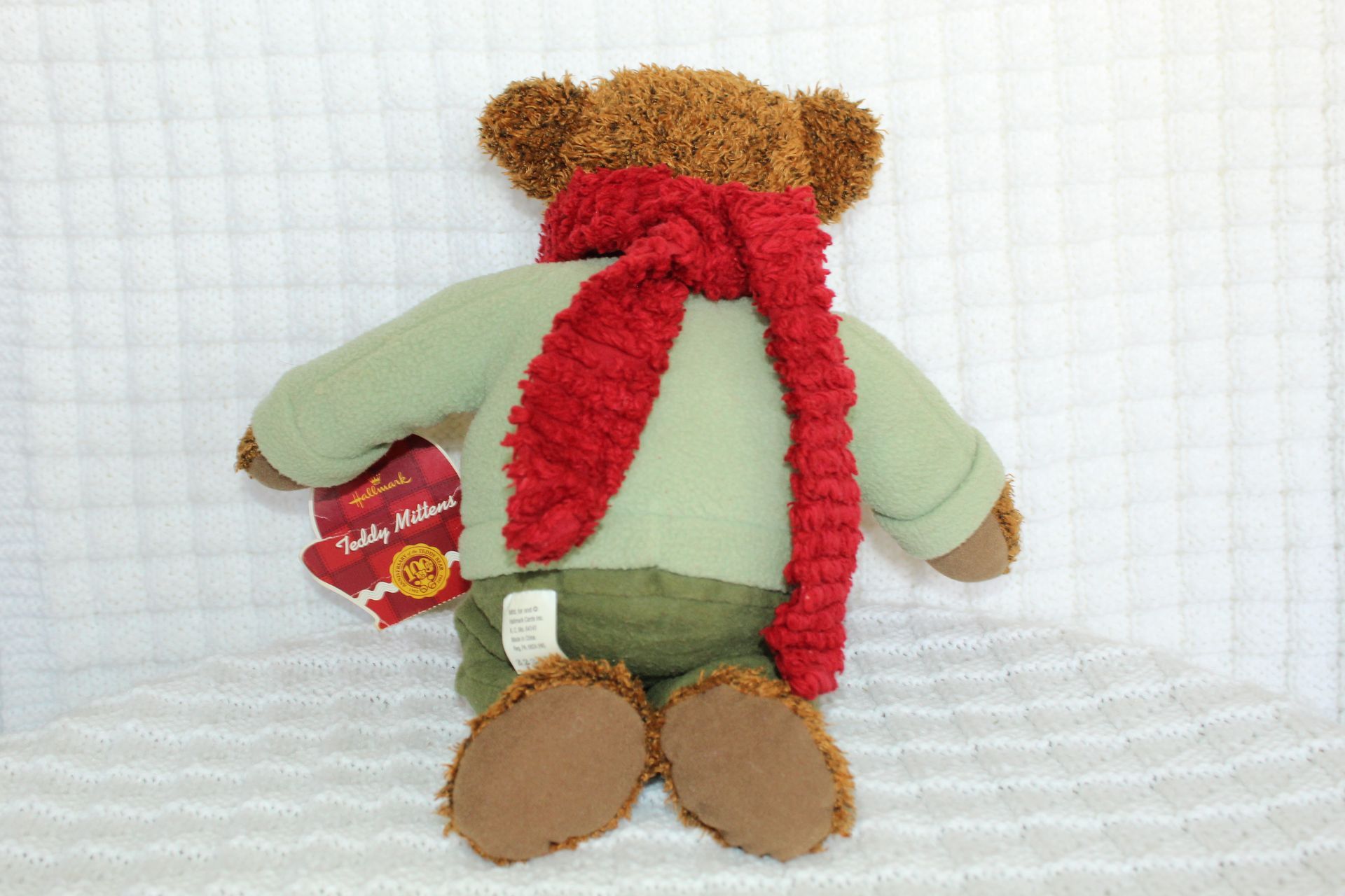 Brown teddy bear in a green sweater and red scarf, sitting beside a red gift tag on a white background