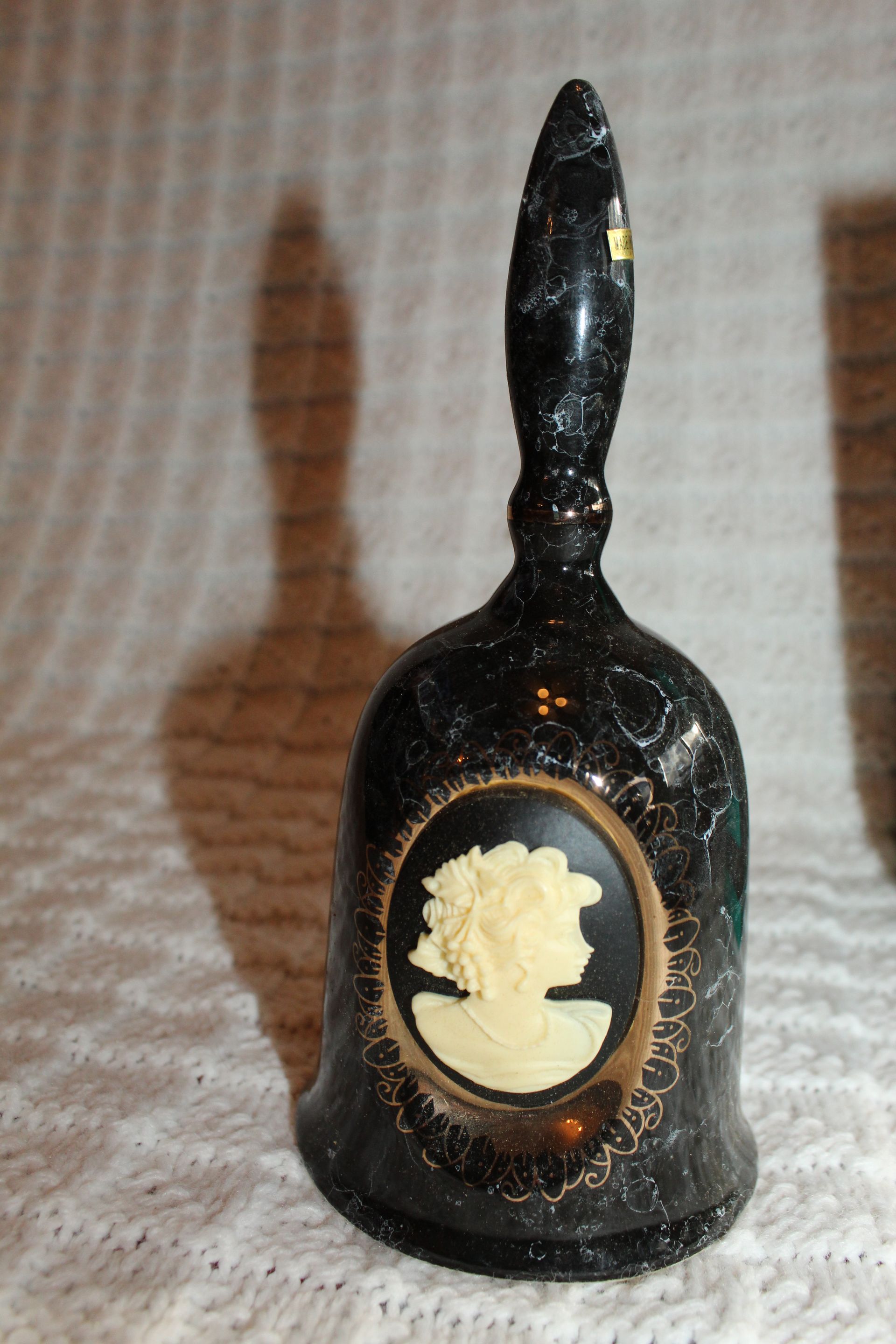 Black decorative bell with cameo portrait on a white textured surface, shadow cast behind it