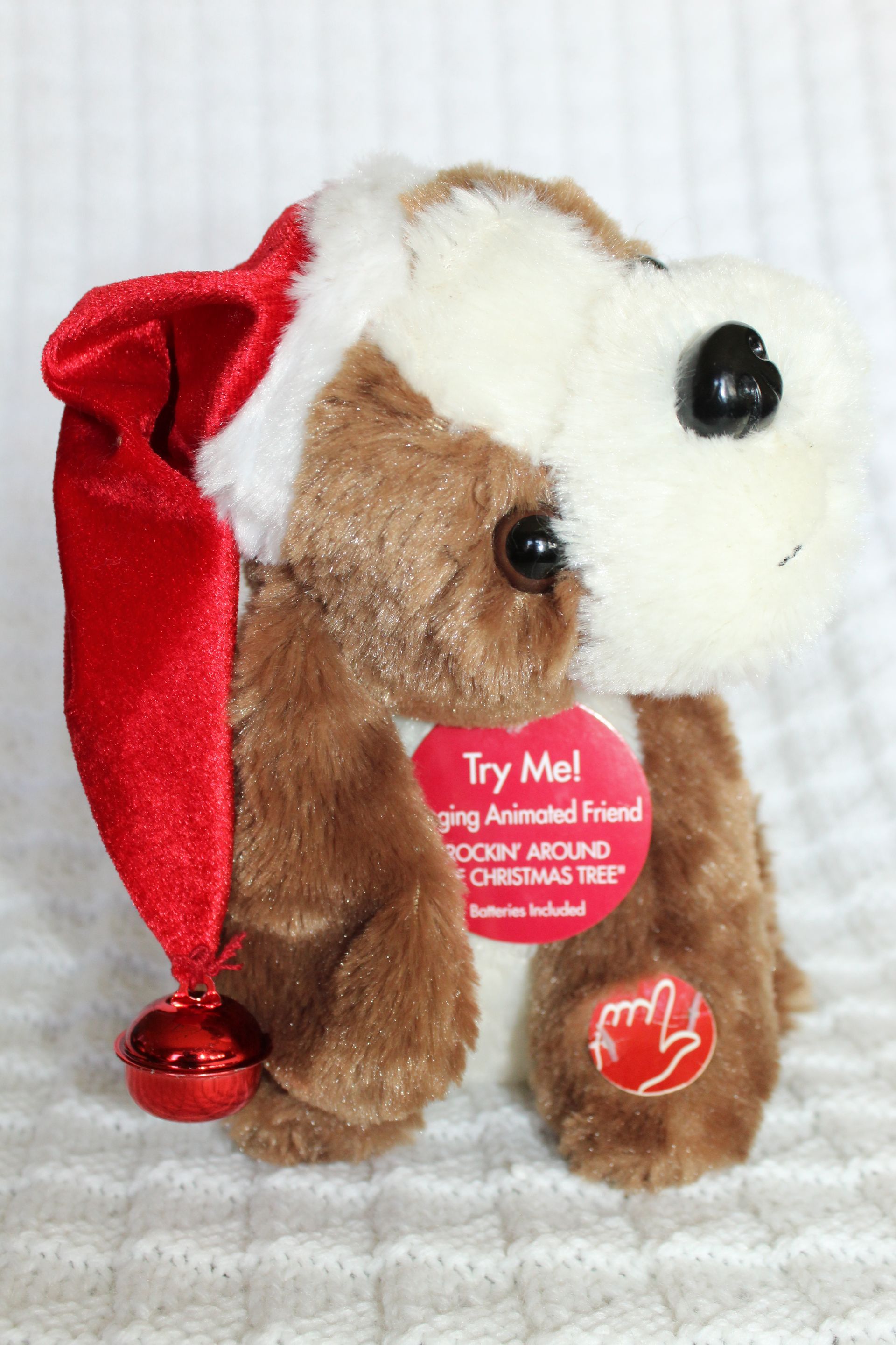 Small brown-and-white plush puppy wearing a red Santa hat and “Try Me” tag