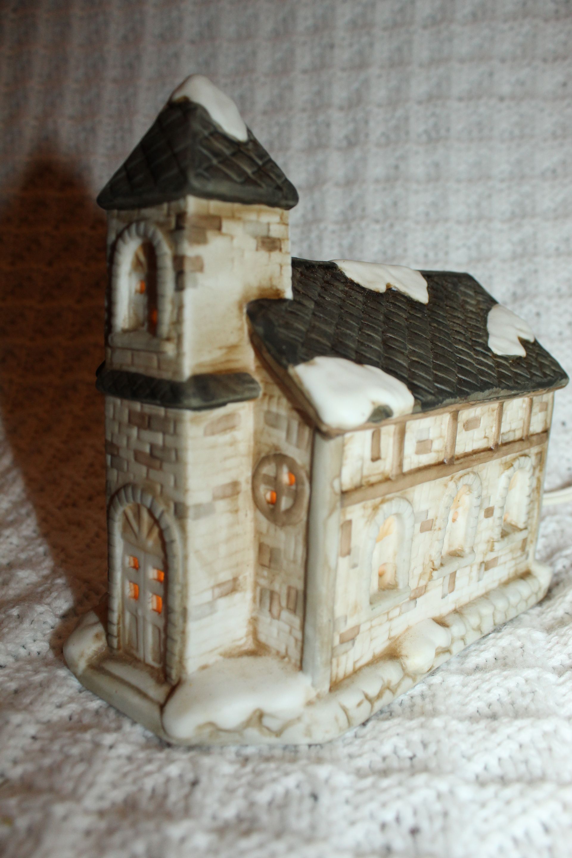 Snow-dusted miniature stone house with a tower, cream walls, and dark roof against a textured white background