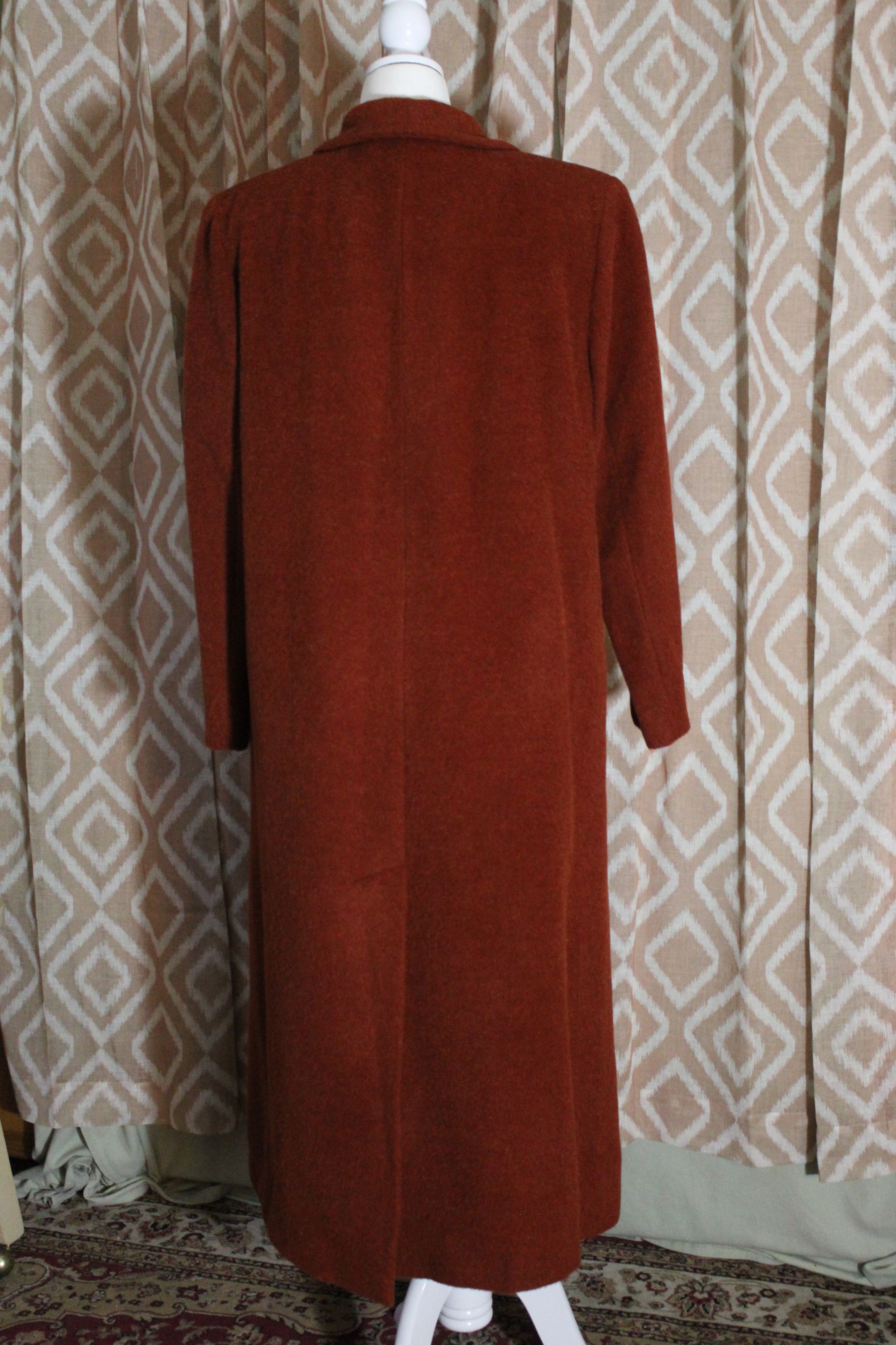 Long rust-colored double-breasted coat on a mannequin against patterned wallpaper.