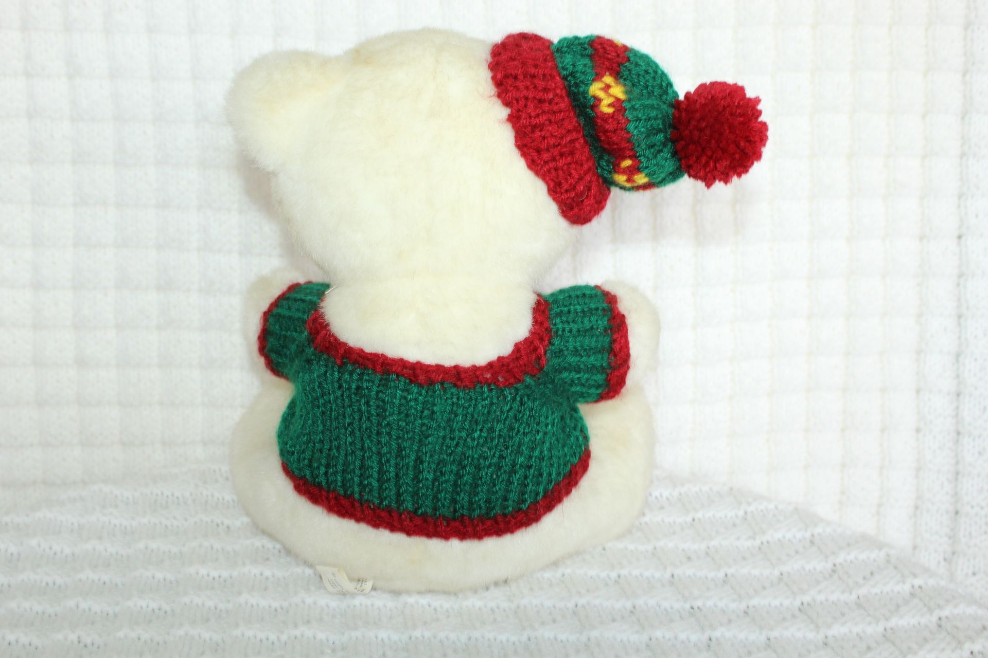 White teddy bear in a red-and-green holiday sweater and hat sitting on a white quilted background