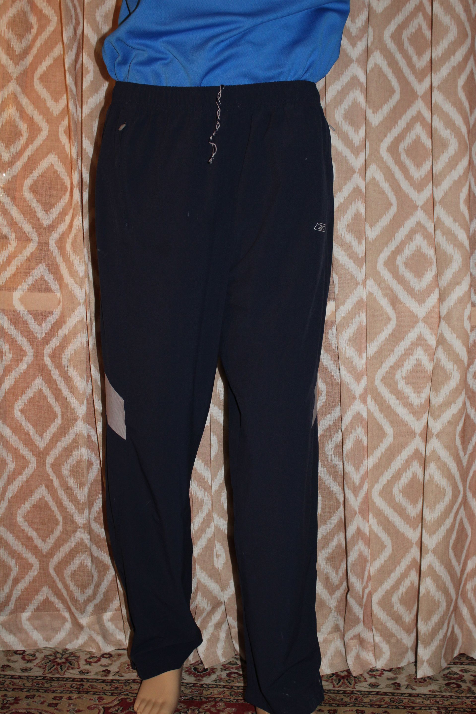 Person standing indoors wearing a blue top and black pants against a patterned curtain.