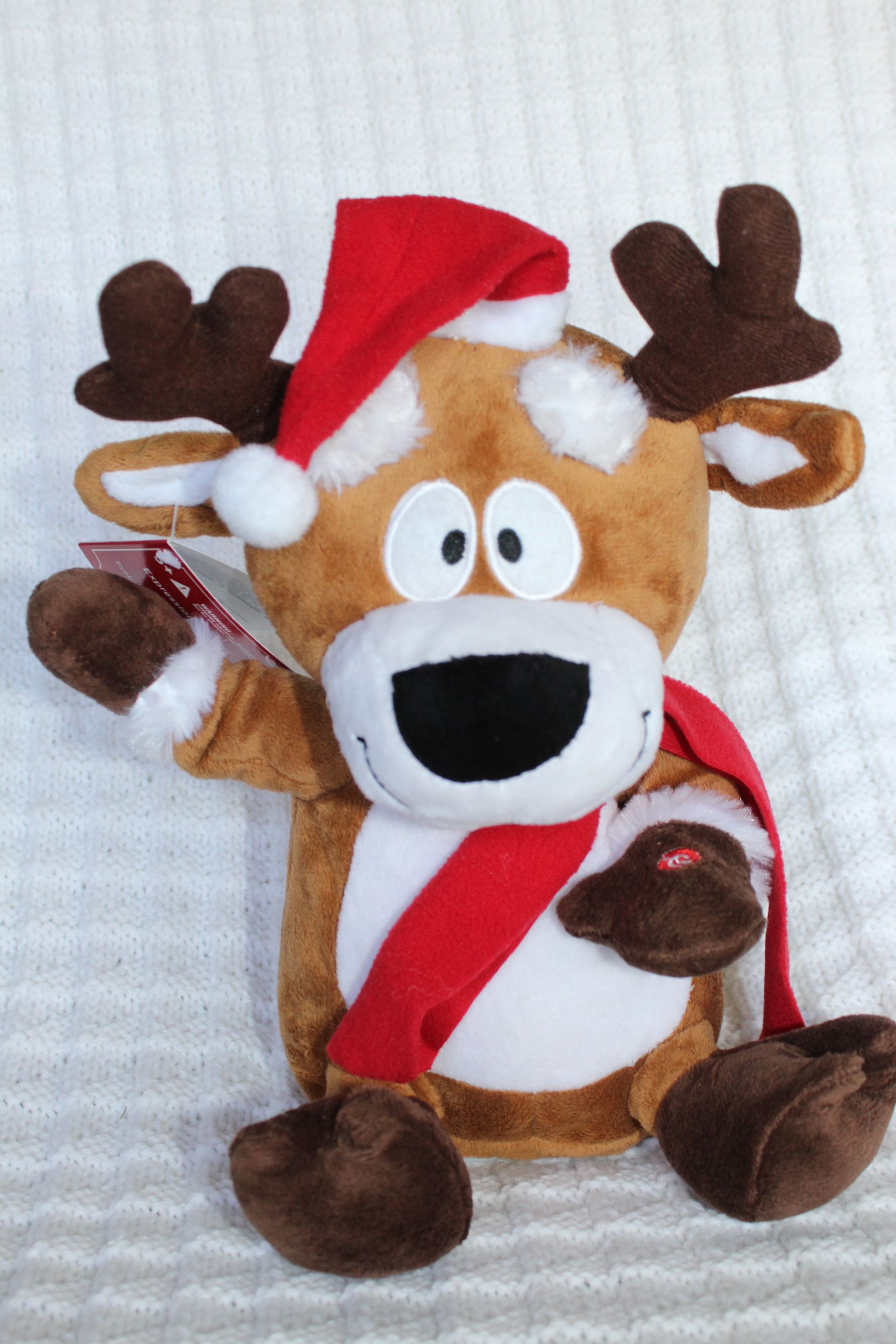 Plush reindeer toy wearing a Santa hat and scarf, sitting on a white textured background.