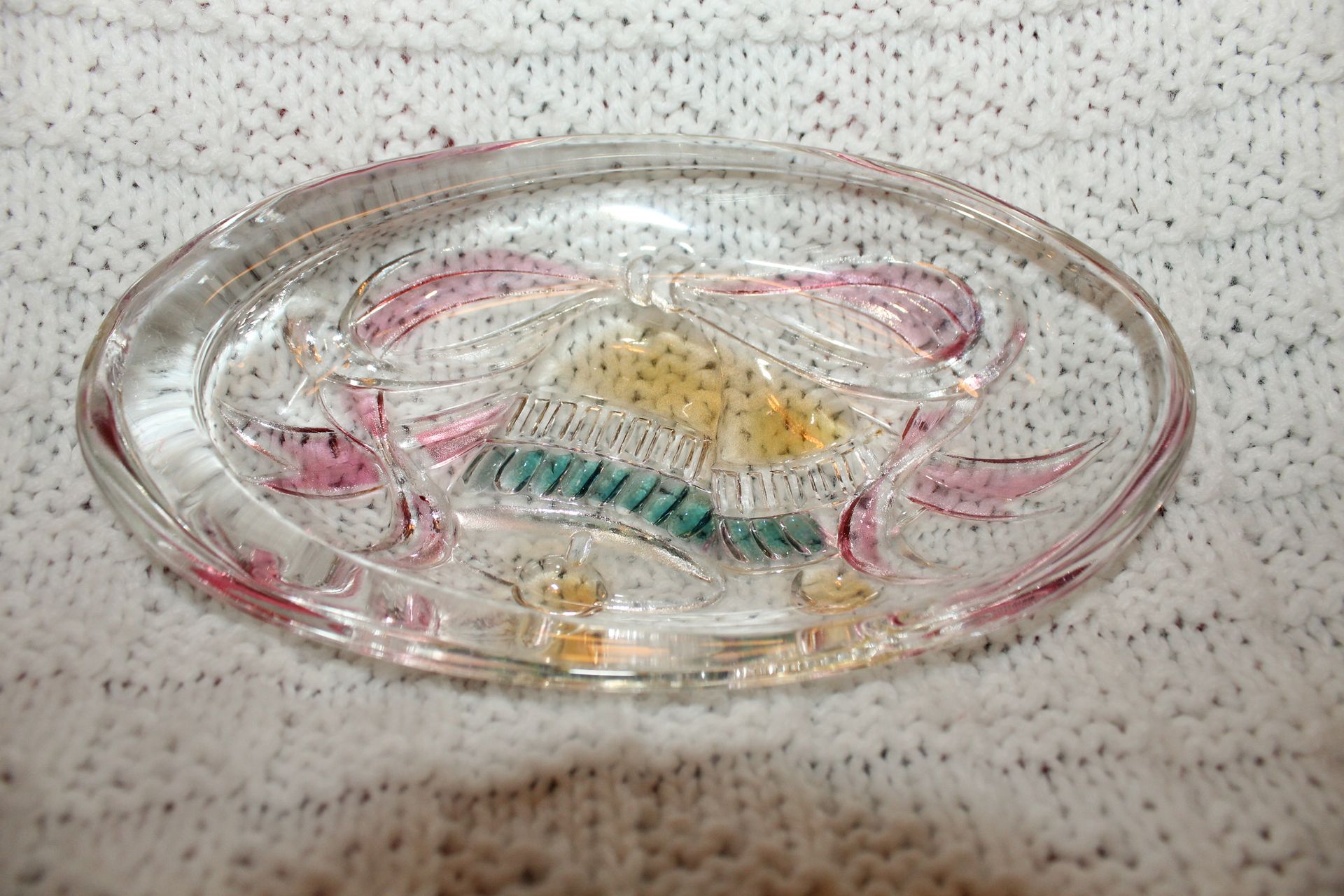 Clear glass oval dish with a colorful fish design on a speckled fabric surface