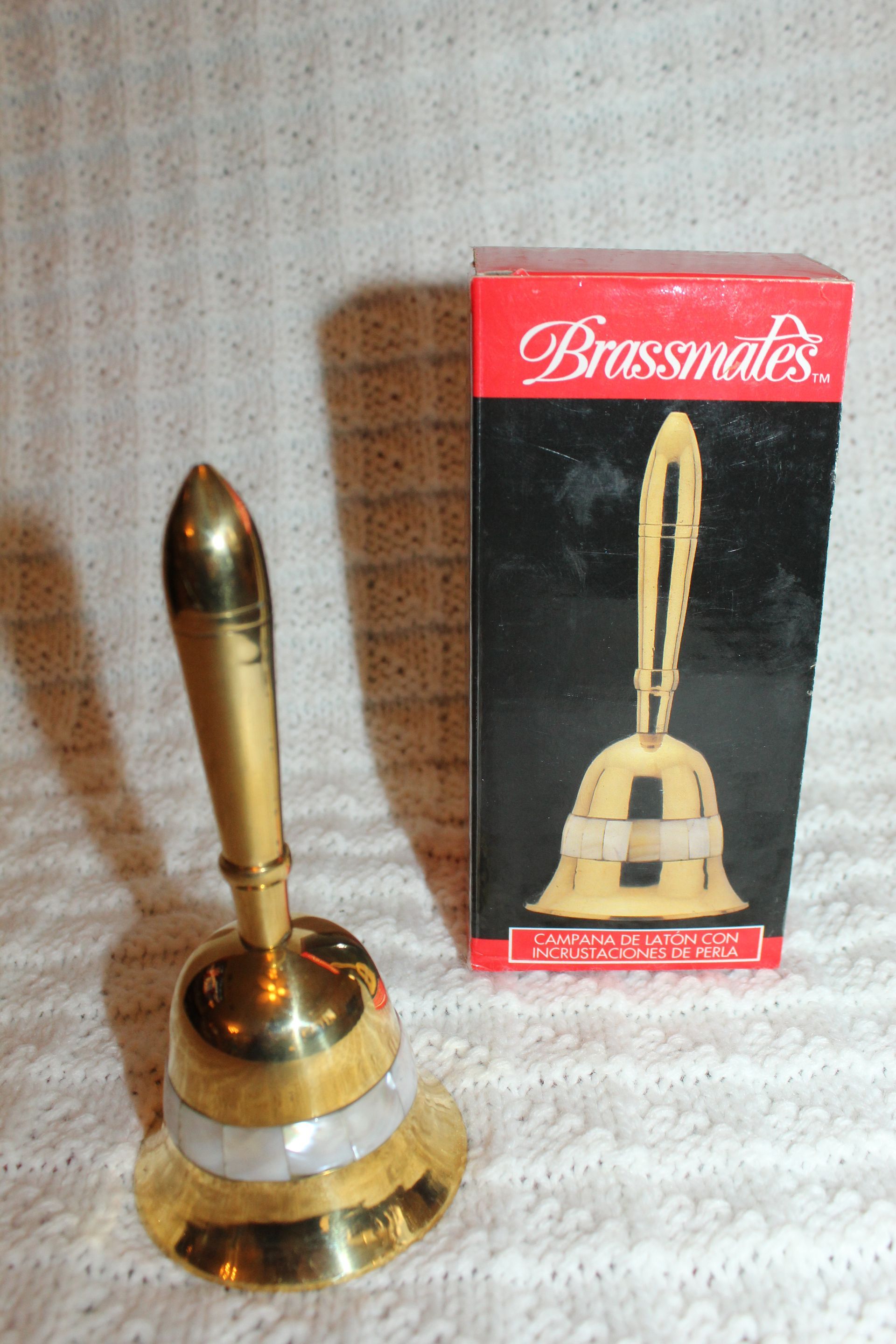 Brass bell with handle beside its red-and-black Brasseal box on a patterned cloth surface