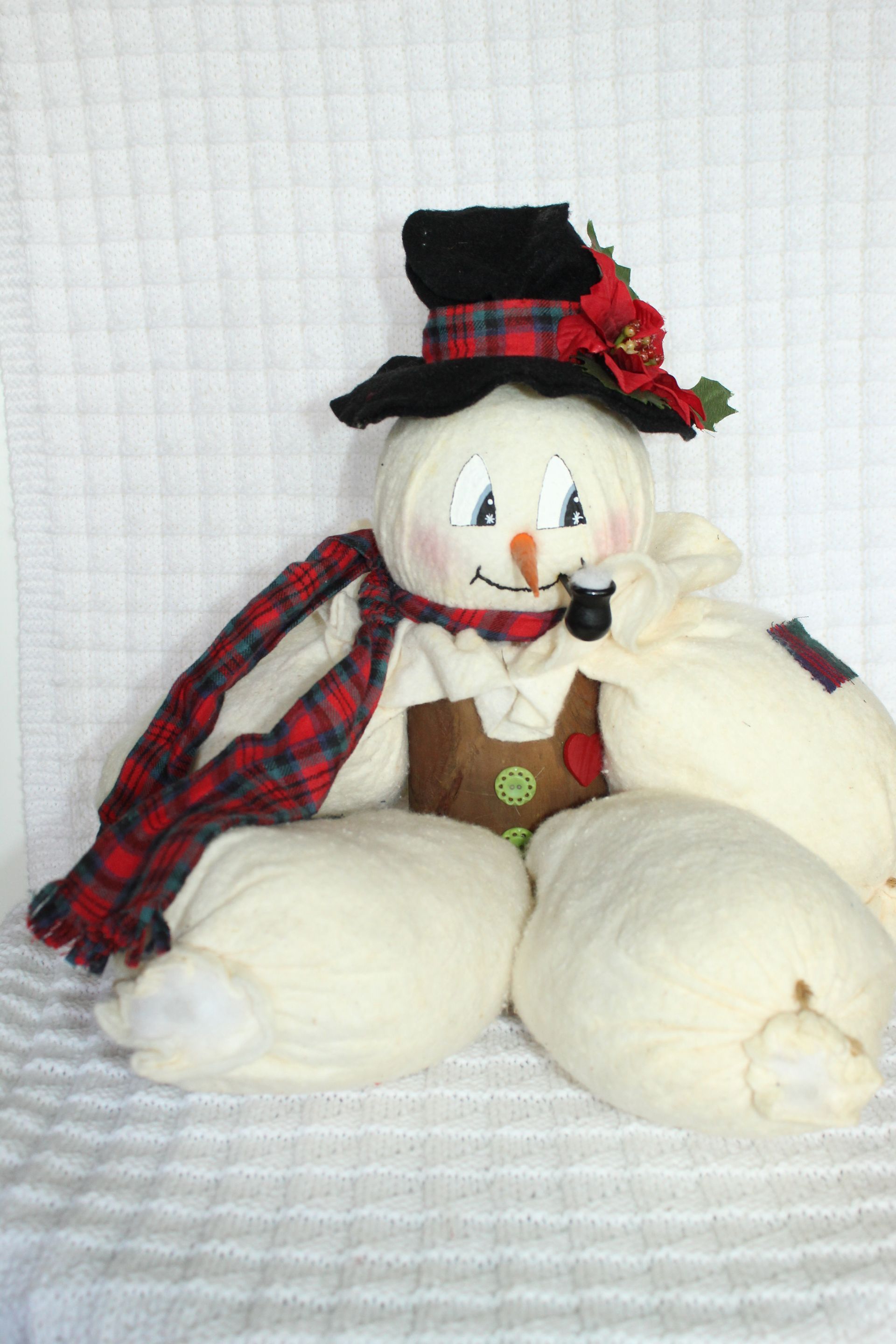 Snowman plush with black hat, red plaid scarf, and flower accent on a white background