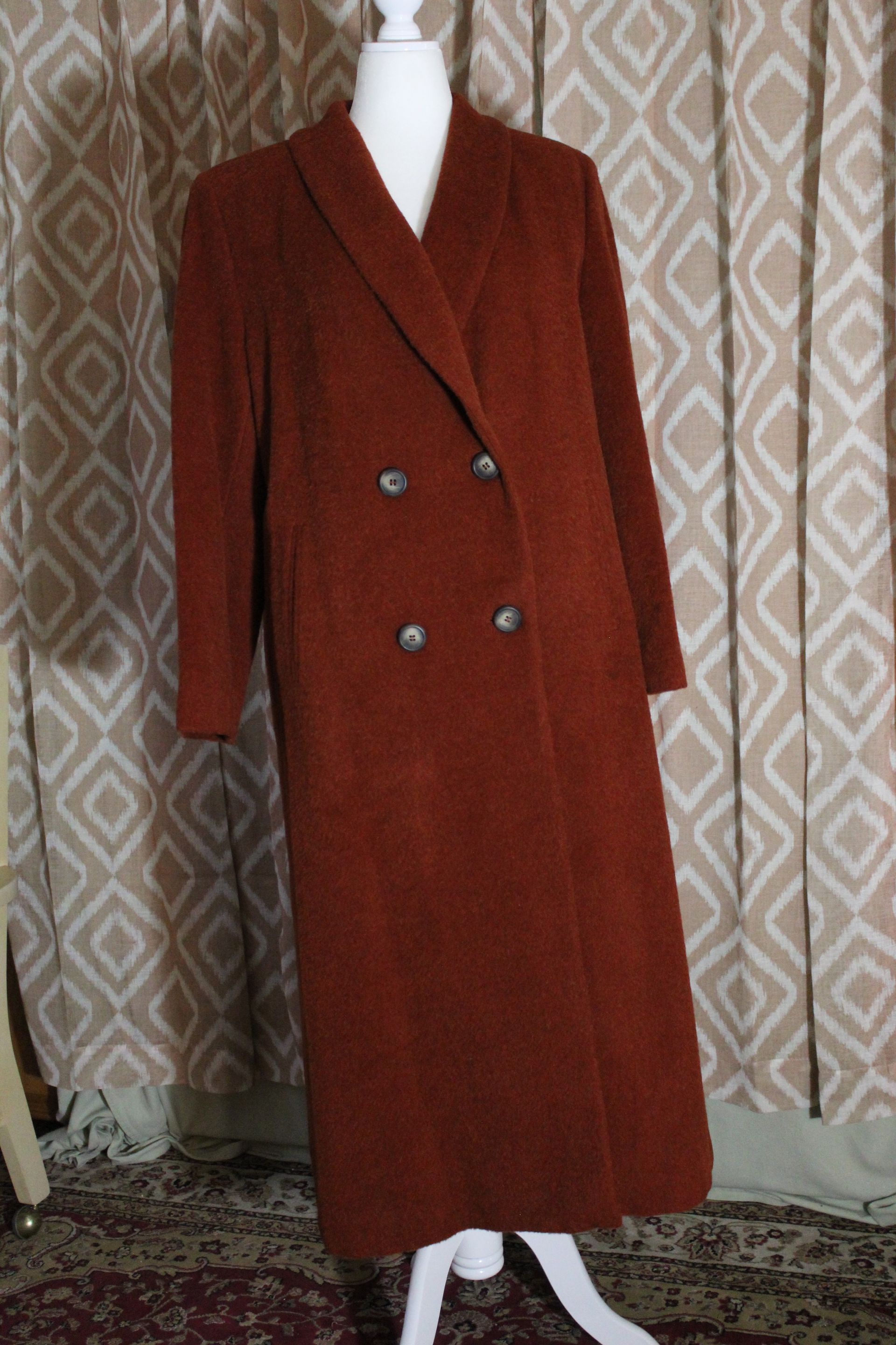 Long rust-colored double-breasted coat on a mannequin against patterned wallpaper.