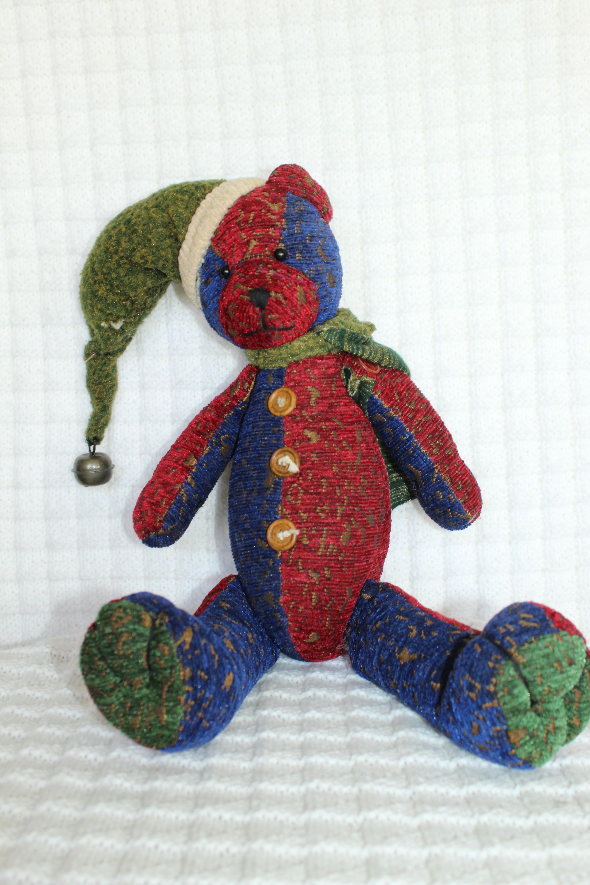 Colorful knitted jester doll with green hat, blue-red body, and dangling bell, sitting on a white surface.