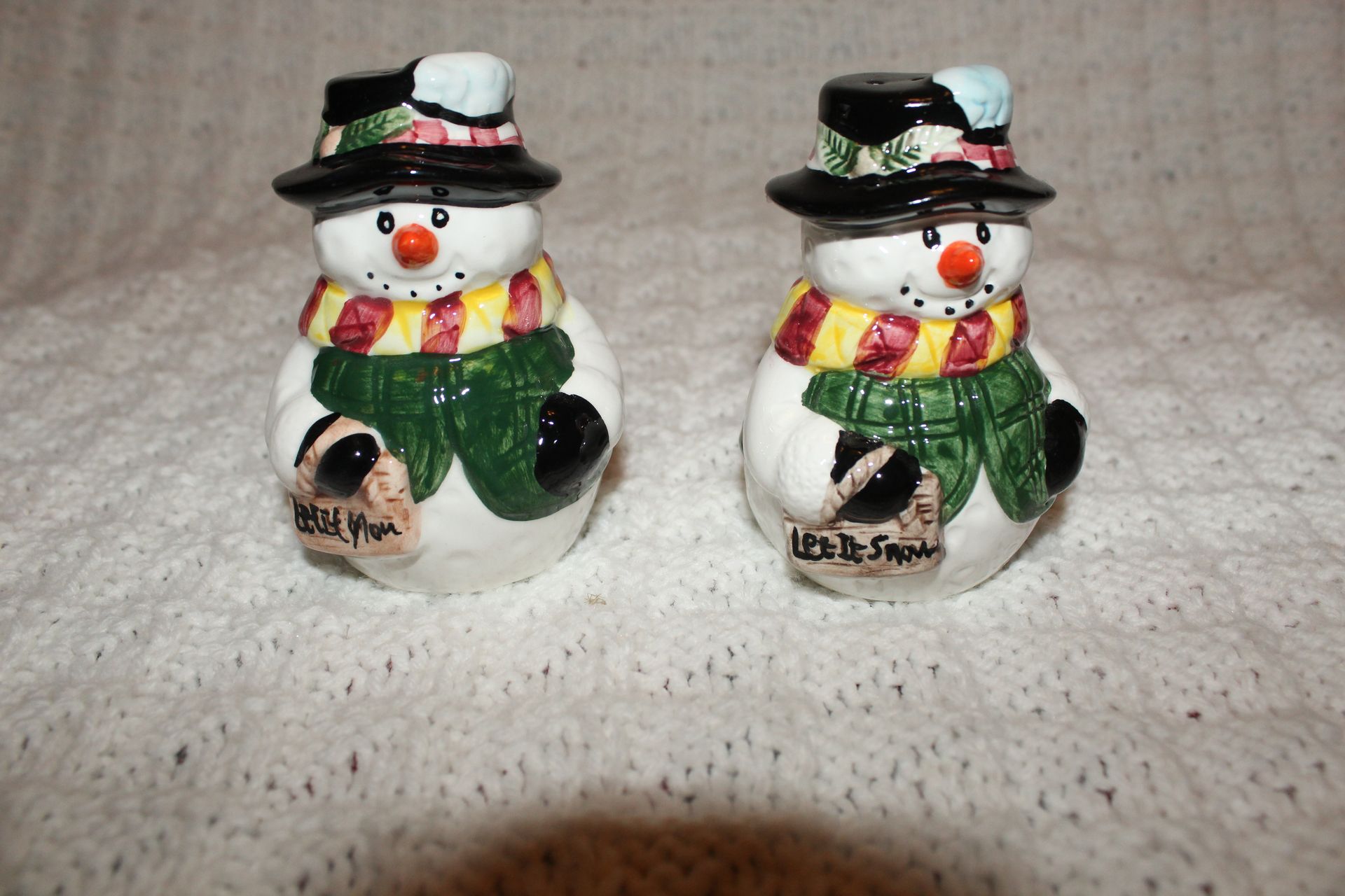 Two small snowman figurines with black hats, colorful scarves, and green mittens on a white blanket