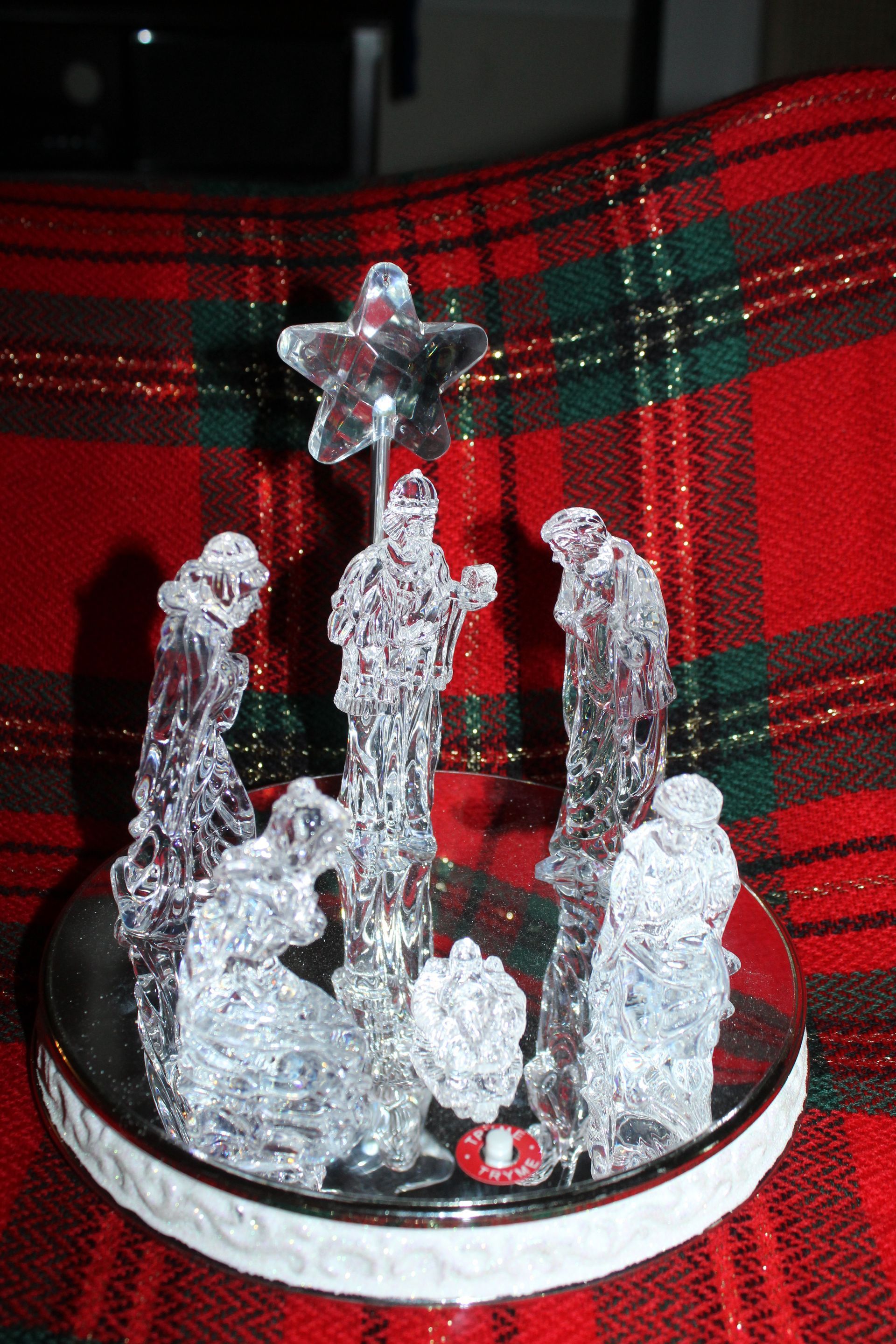 Small clear nativity figurines on a round white base against red-and-green plaid fabric.