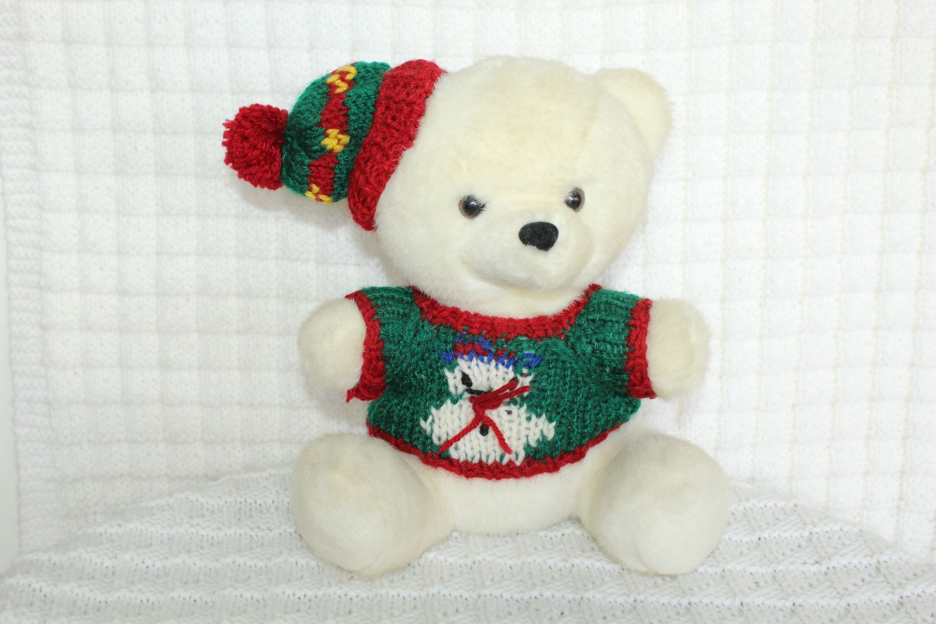 White teddy bear in a red-and-green holiday sweater and hat sitting on a white quilted background