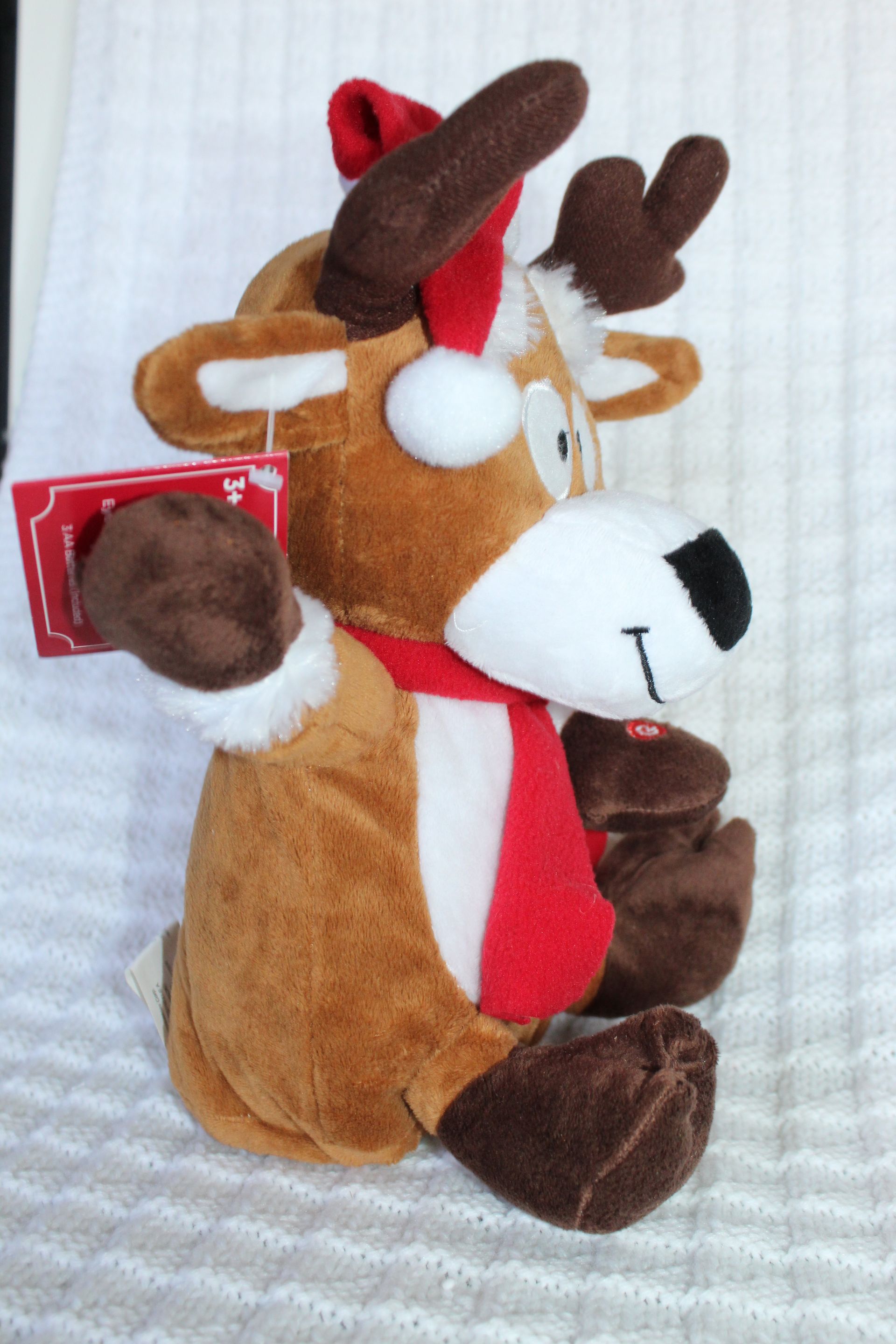 Plush reindeer toy wearing a Santa hat and scarf, sitting on a white textured background.