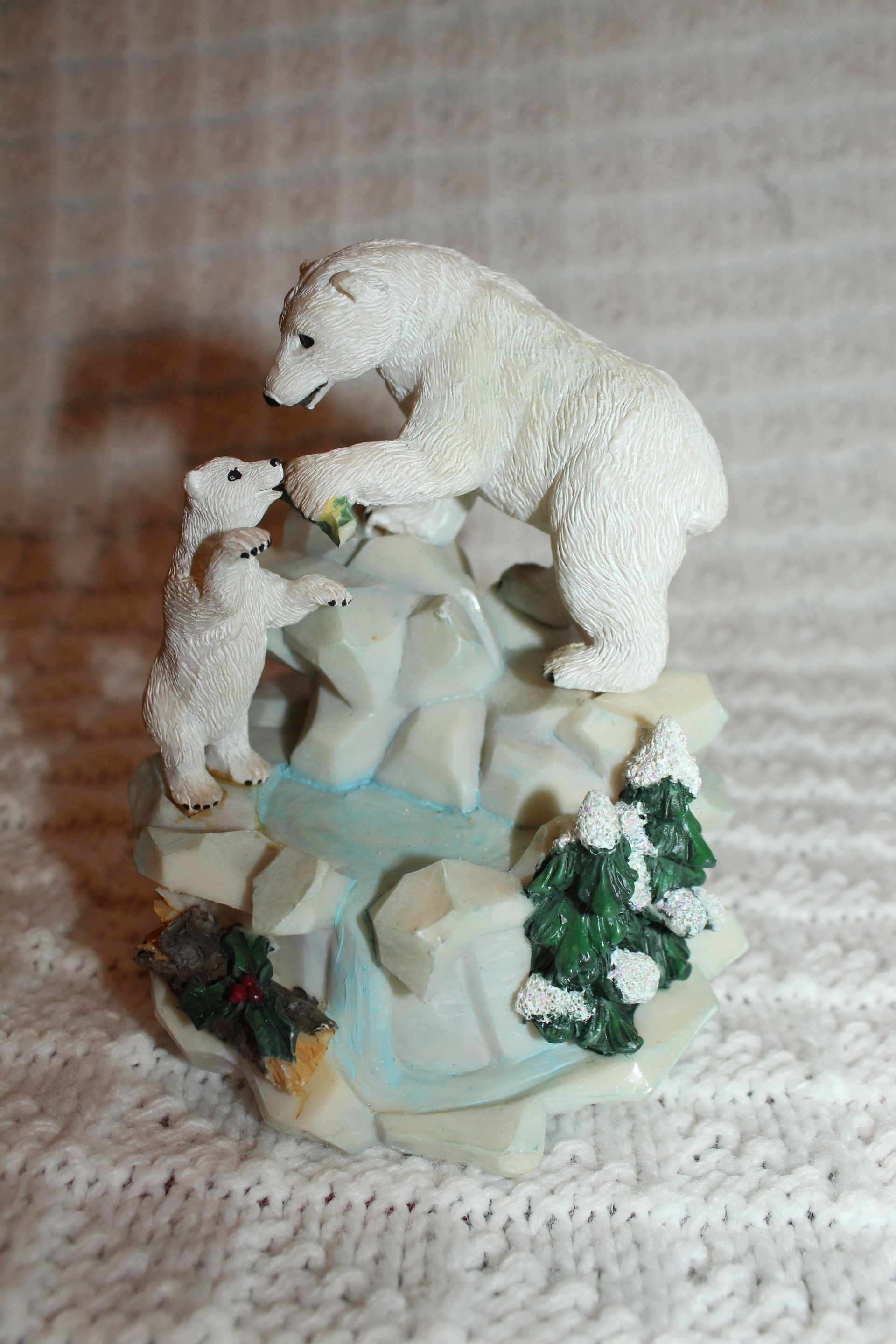 Small white bear figurine on a rocky base, reaching toward a smaller bear amid green foliage.