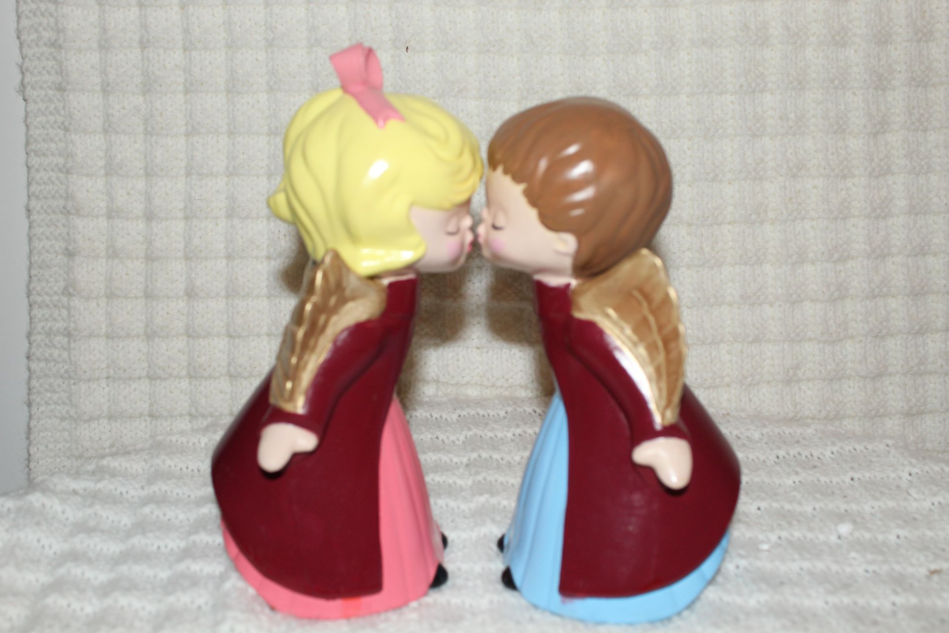 Two small figurines facing and touching noses, one blonde and one brown-haired, in red dresses.