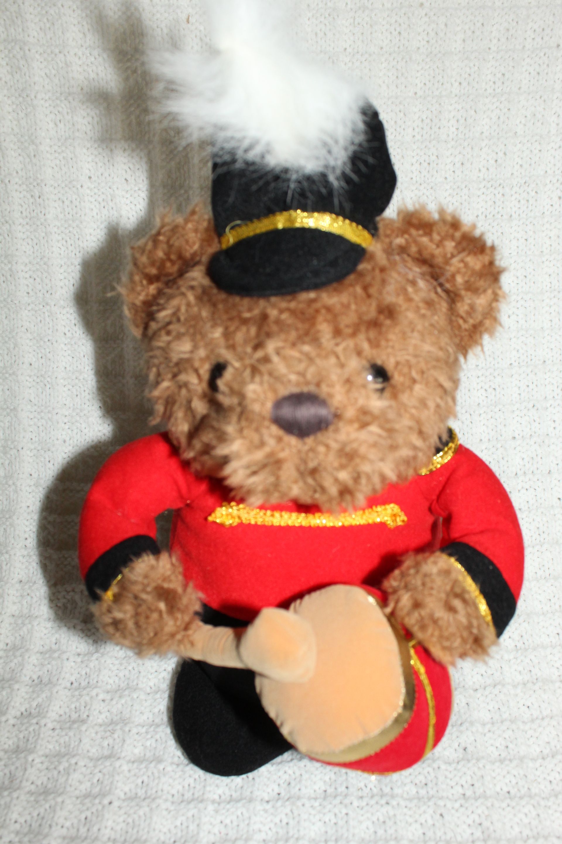 Brown teddy bear in red outfit holding a drum, with a black-and-white feathered hat