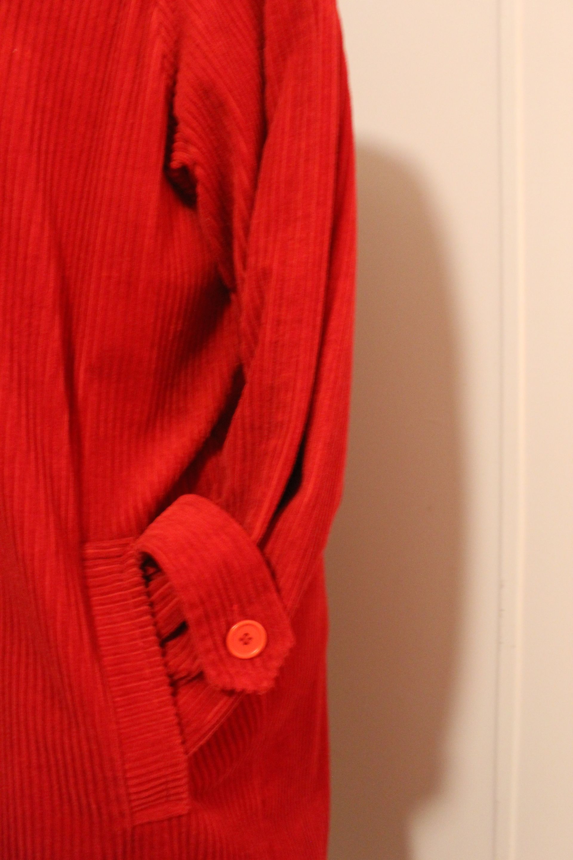 Red ribbed sweater sleeve with a buttoned cuff against a beige wall.