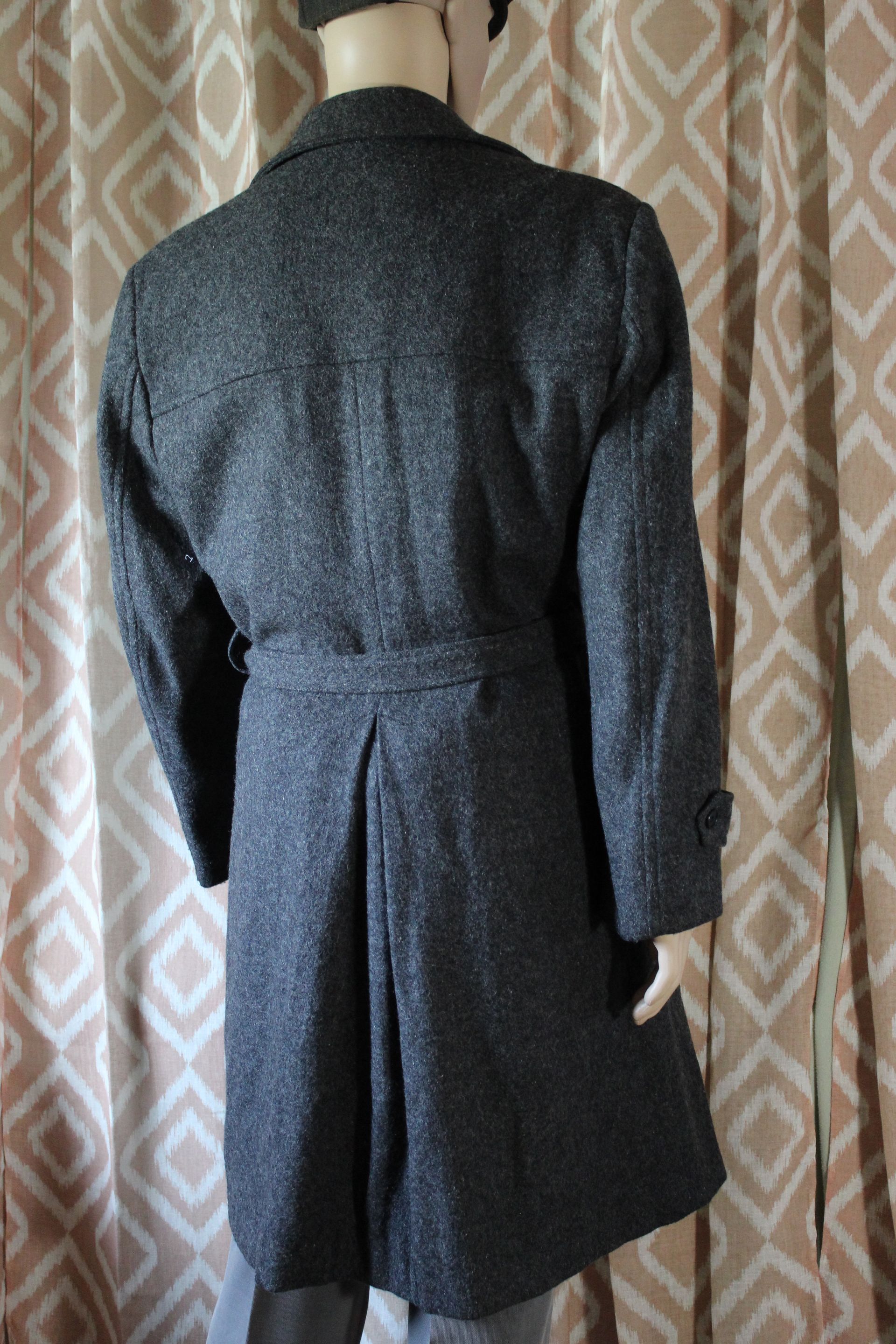 Mannequin wearing a dark gray overcoat, black top, and gray trousers against a patterned curtain