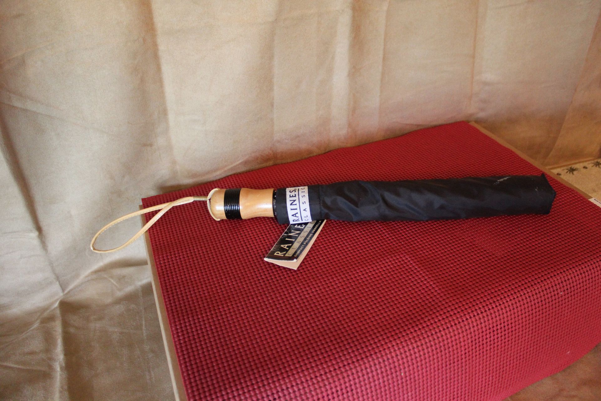 Close-up of a black Raines compact folding umbrella with tan handle and gold label on red fabric background