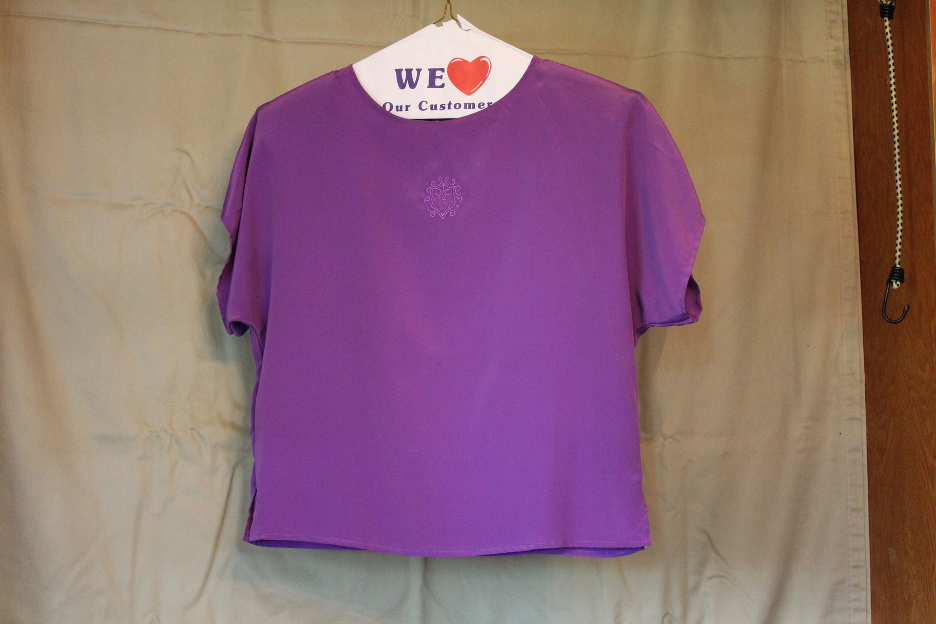 Purple short-sleeve T-shirt hanging against a beige backdrop with a white heart tag at the collar