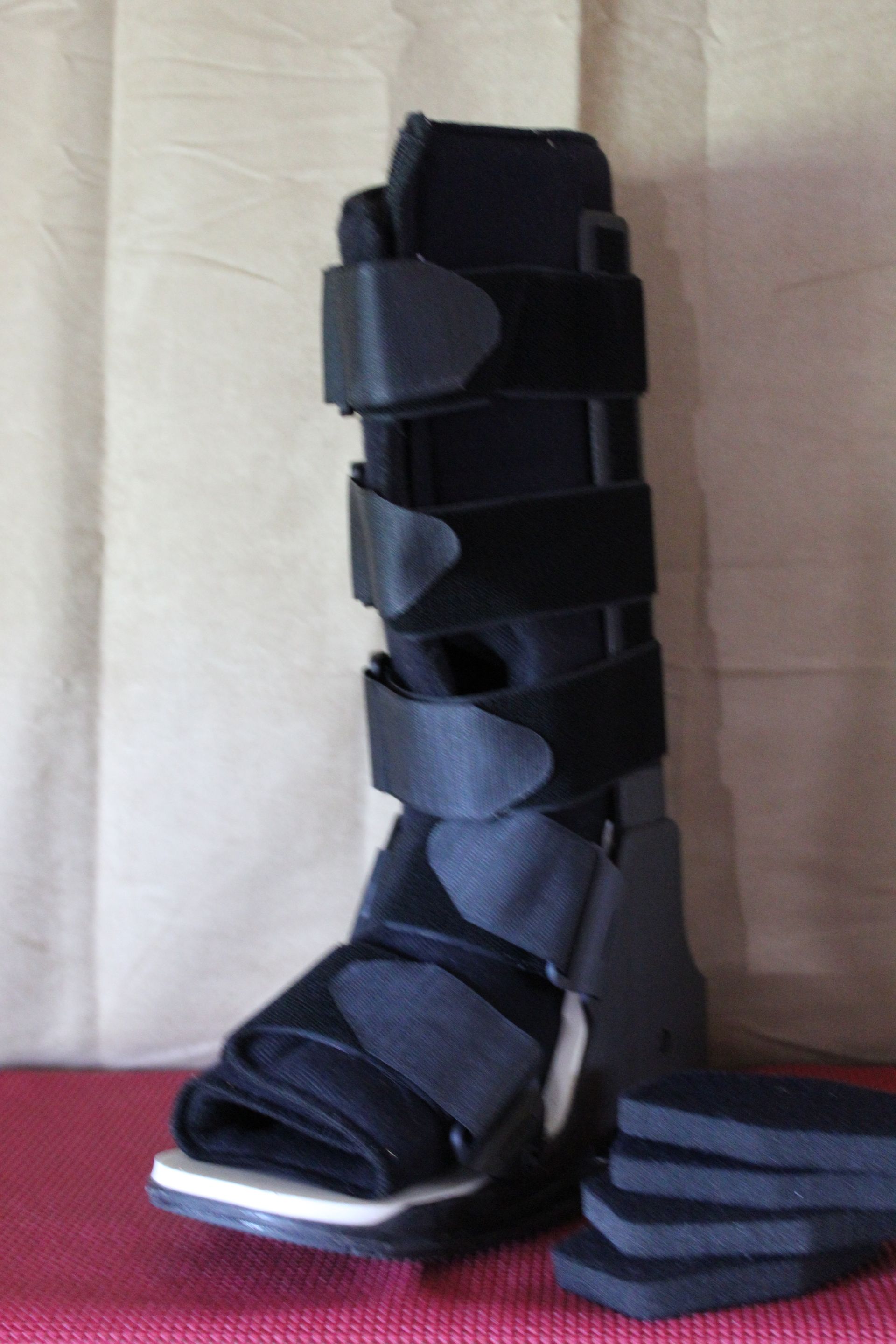 Black orthopedic walking boot with straps, shown upright next to stacked inserts on a red surface.