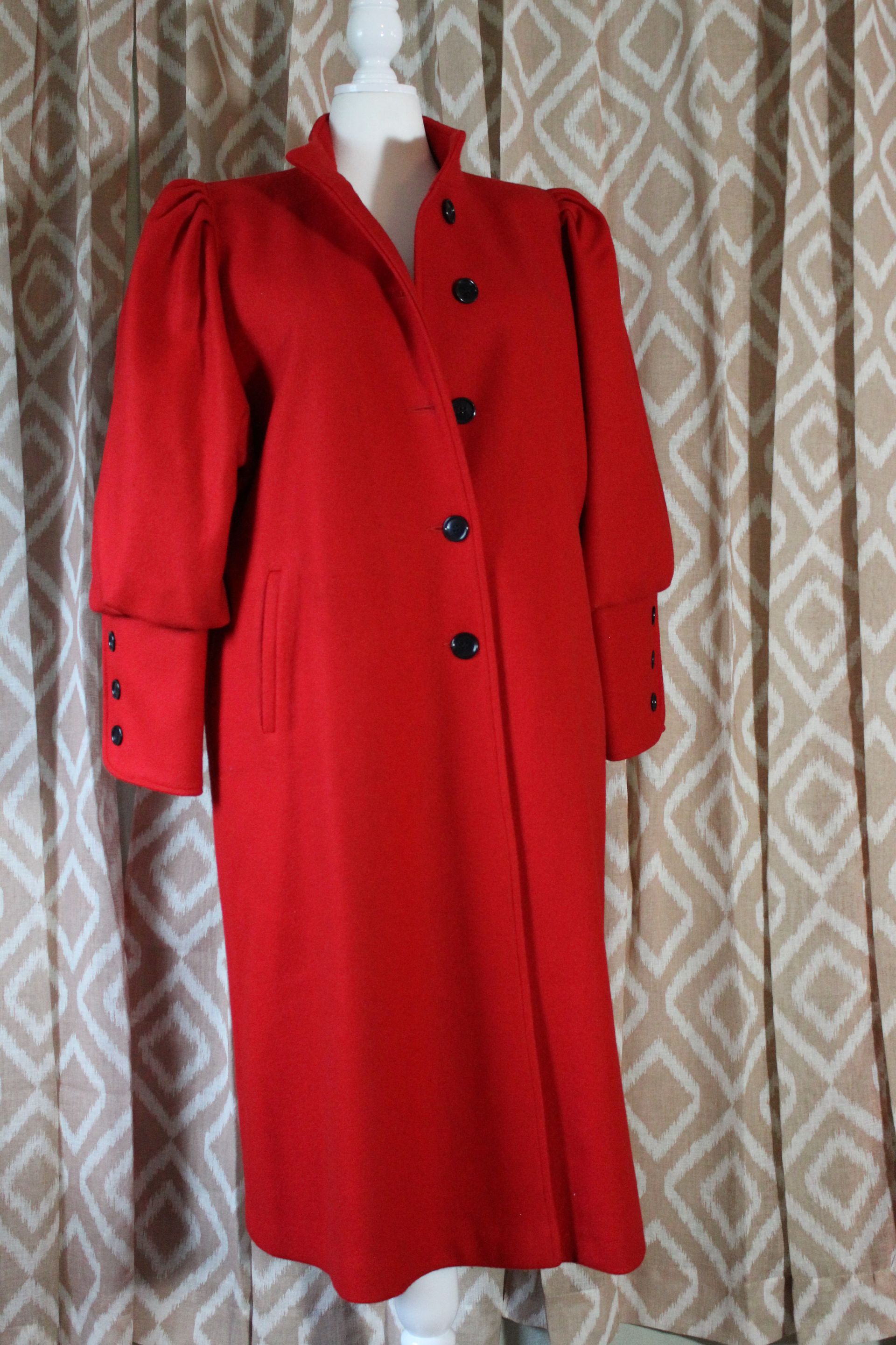 Red knee-length coat with black buttons on a mannequin against a patterned curtain backdrop