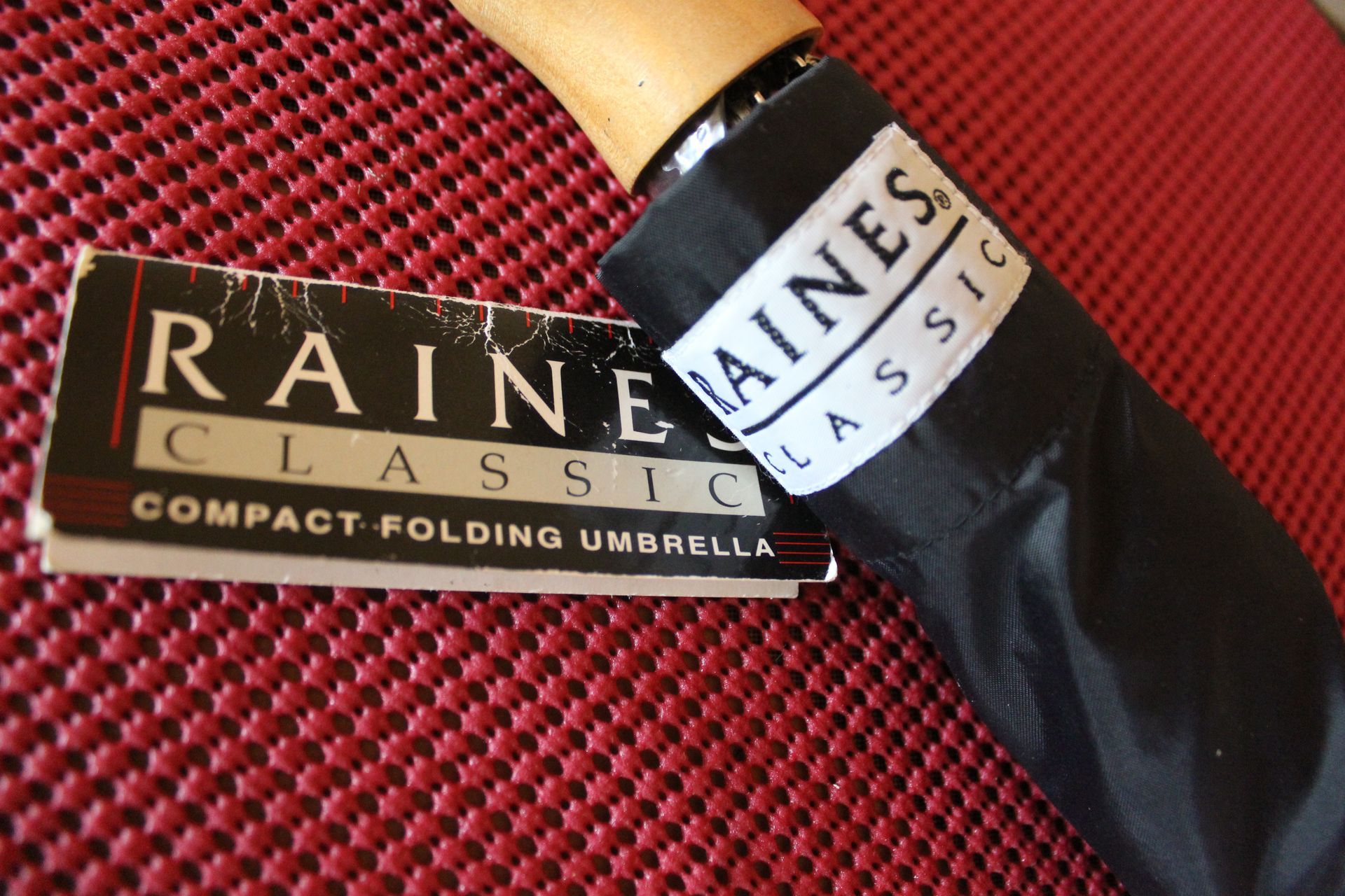 Close-up of a black Raines compact folding umbrella with tan handle and gold label on red fabric background