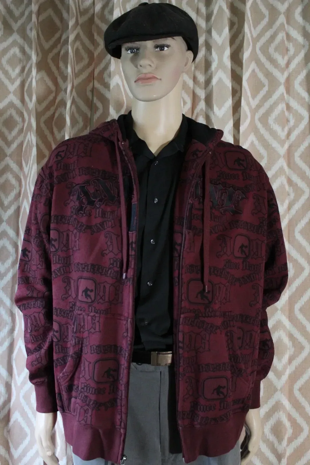 Mannequin wearing a black cap, black shirt, and maroon patterned jacket against a patterned curtain backdrop