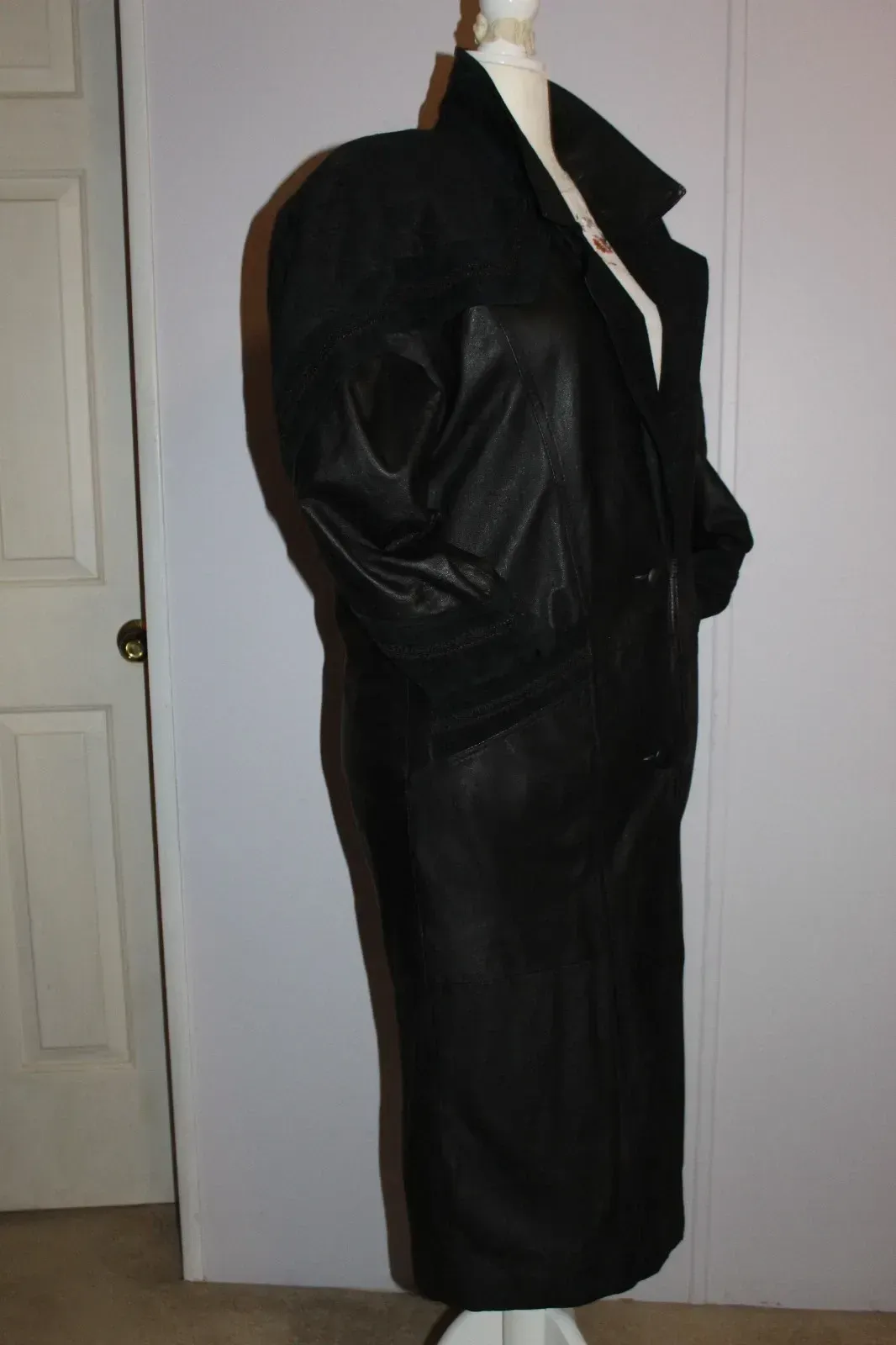 Black leather coat on a mannequin against a wall, shown in side view.