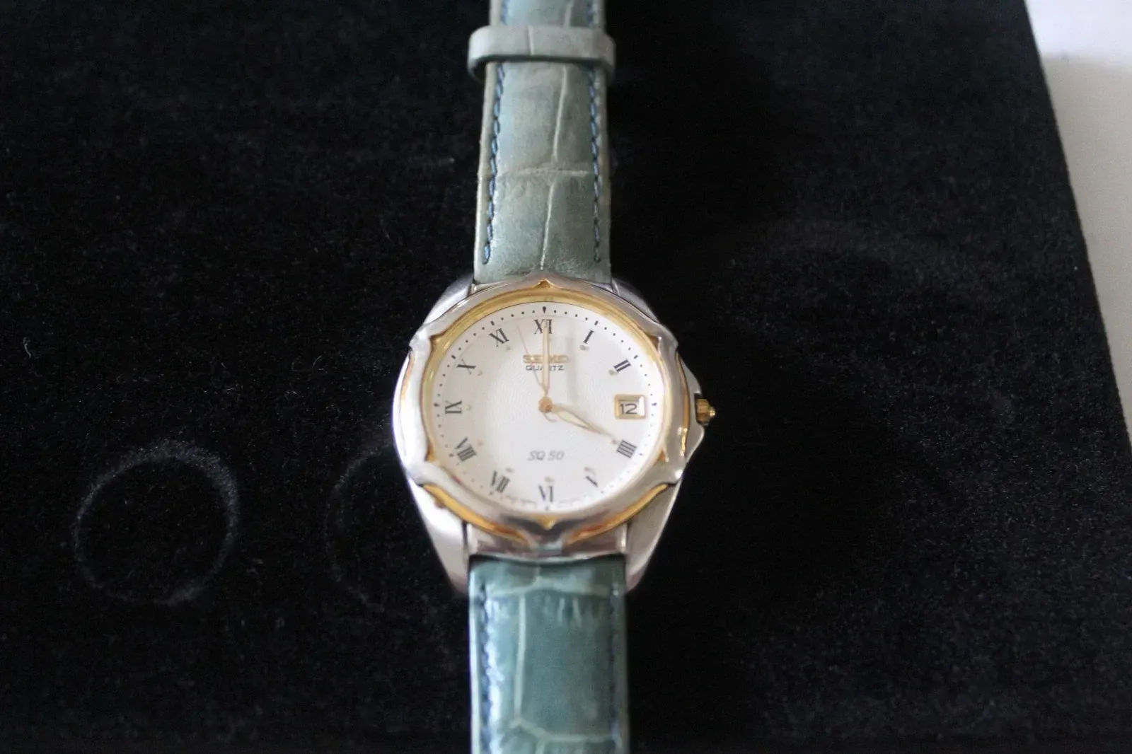 Gold watch with white face and green leather strap on a dark background