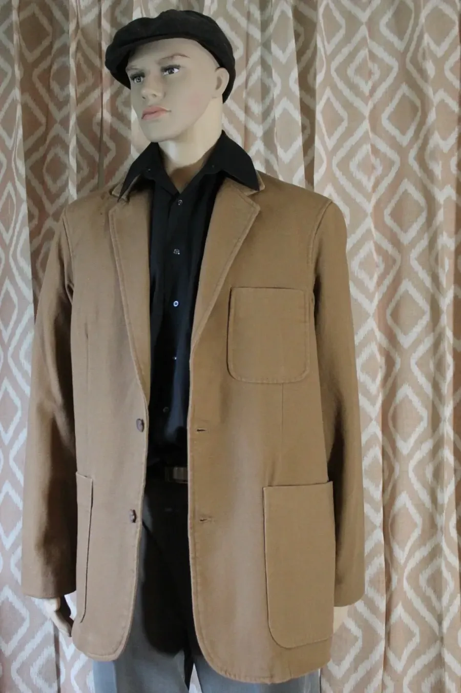 Person wearing a tan blazer and black hat stands in front of patterned curtains.
