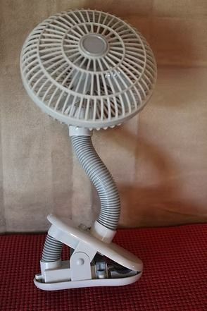 White clip-on desk fan with a flexible neck against a brown wall and red surface.