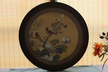Decorative round wall art with birds and flowers in a dark frame, set against bamboo blinds