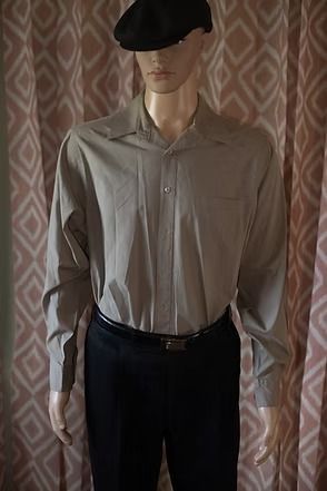 Mannequin wearing a beige button-up shirt, dark pants, and a black cap in front of patterned curtains