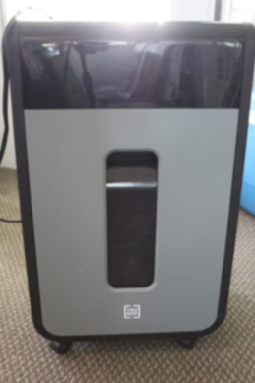 Black and gray office shredder with a narrow paper slot, standing on a carpeted floor.