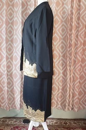 Black jacket and skirt with gold trim displayed on a mannequin against patterned curtains