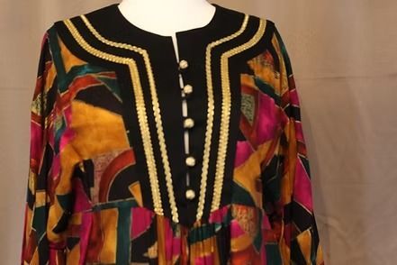 Colorful geometric-patterned long dress with black yoke and gold trim on a mannequin, against a curtain backdrop