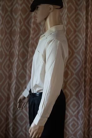 Mannequin wearing a cream button-up shirt, black pants, and a black hat against patterned wallpaper.