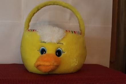 Yellow plush basket with a handle and duck face, with white stuffing on top, sitting on a red surface.