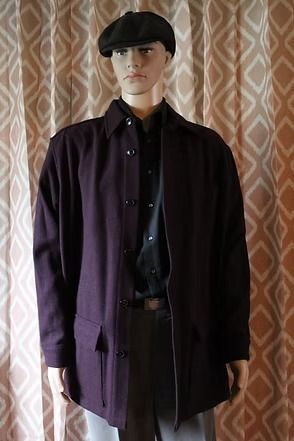 Person standing indoors wearing a black beret, dark shirt, and purple jacket in front of patterned curtains