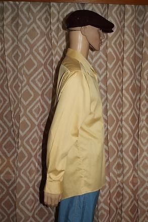 Mannequin in a yellow shirt, blue pants, and black hat standing before patterned curtains