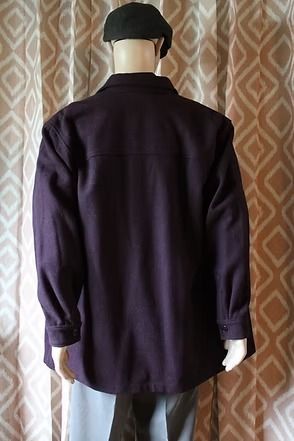 Person standing indoors wearing a black beret, dark shirt, and purple jacket in front of patterned curtains