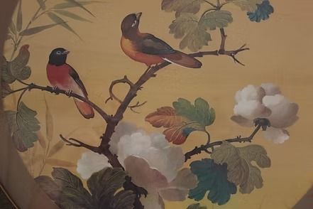Decorative round wall art with birds and flowers in a dark frame, set against bamboo blinds