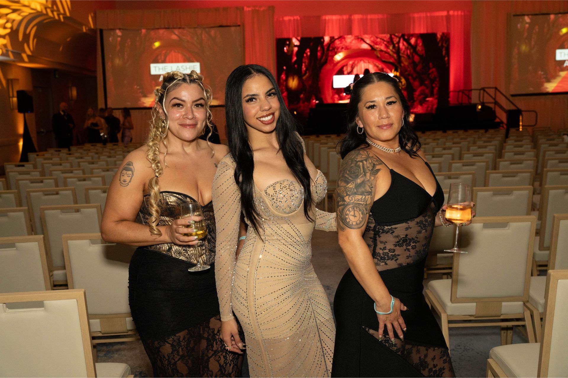 Three women in formal dresses standing together in a banquet hall with rows of chairs and red stage lighting.