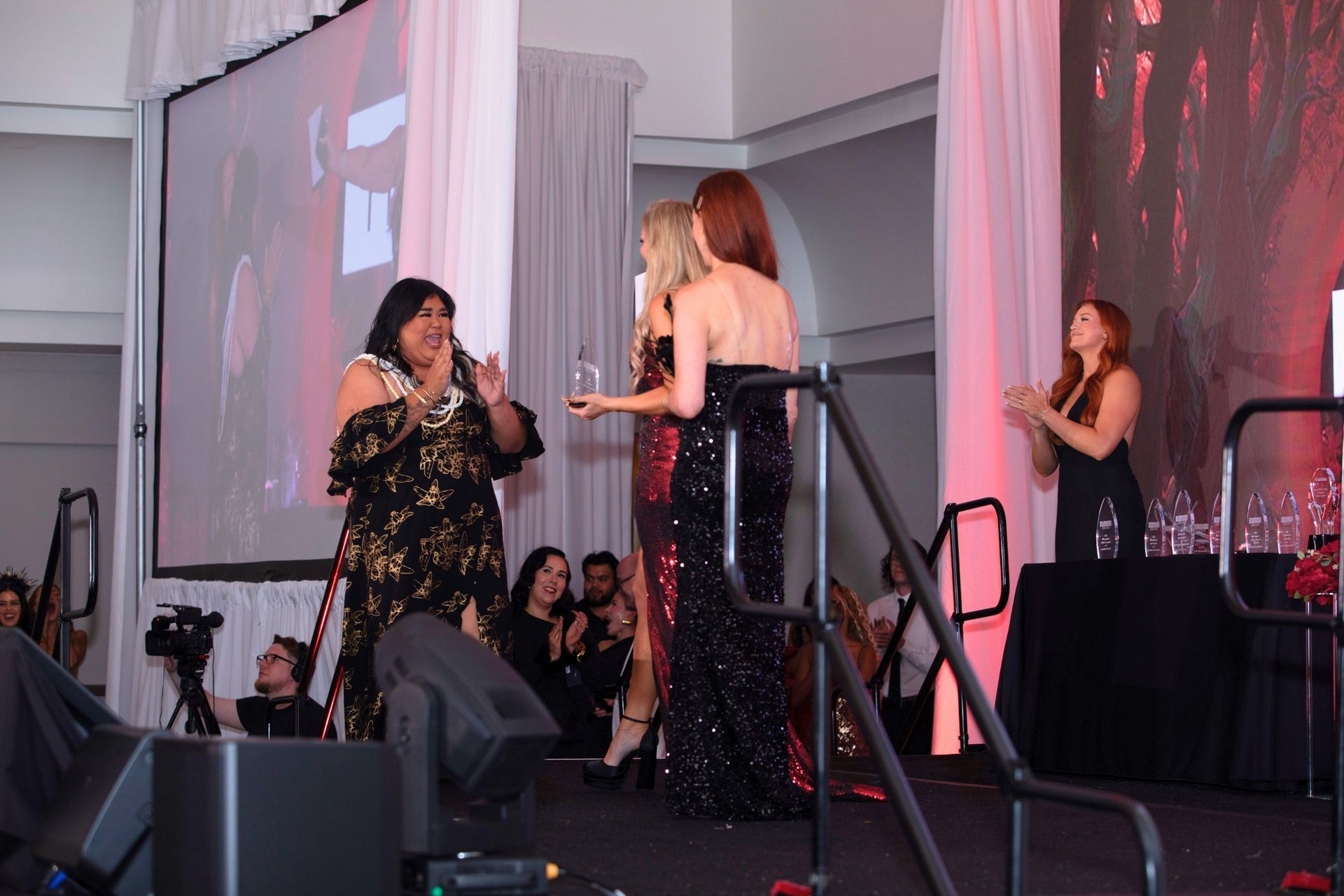 Women on a stage at a formal event, one holding an award while others applaud nearby.