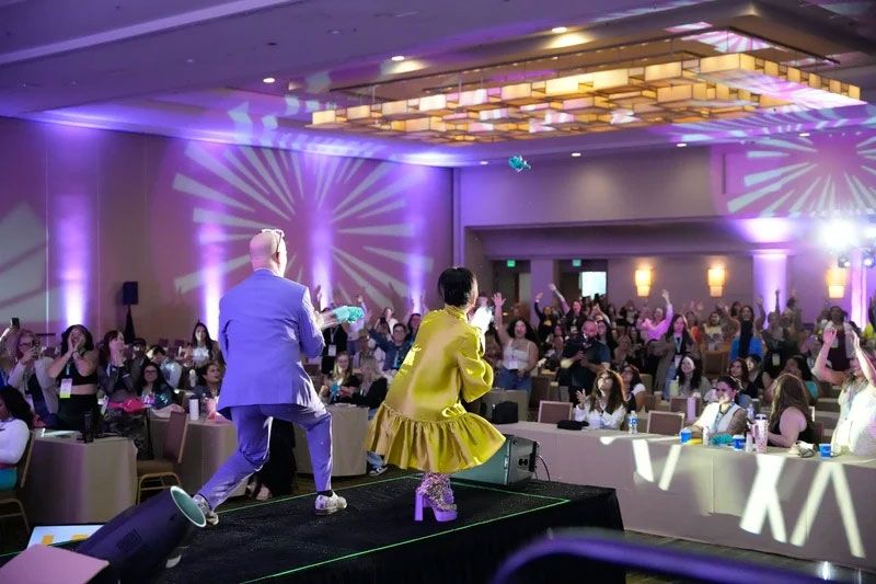 Two people on stage in front of a crowd; spotlights and projections in a ballroom.
