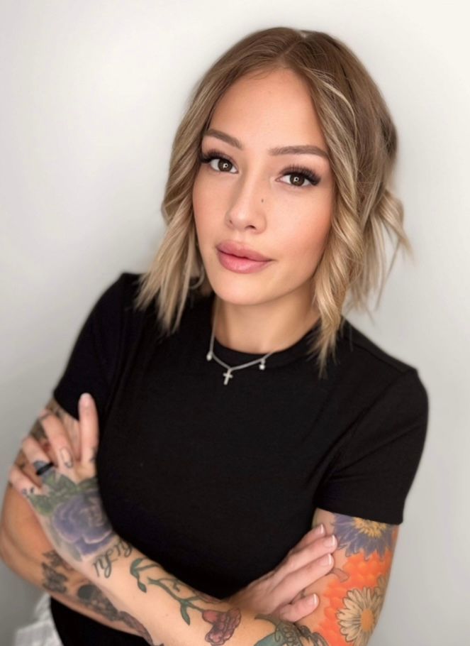 Person with tattoos and blonde hair in a black shirt, posing with arms crossed against a plain background