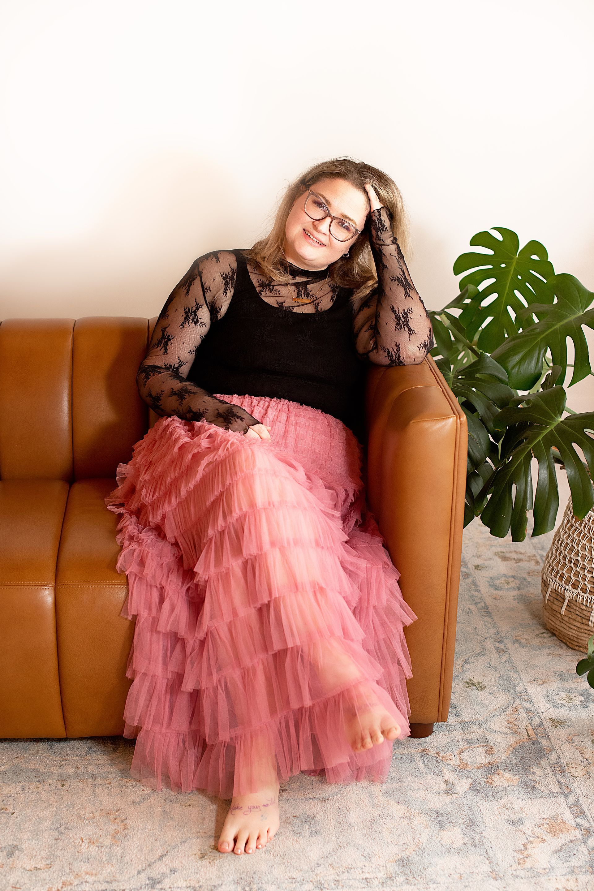 Woman reclining on a tan sofa in a black lace top and pink ruffled skirt beside a potted plant.