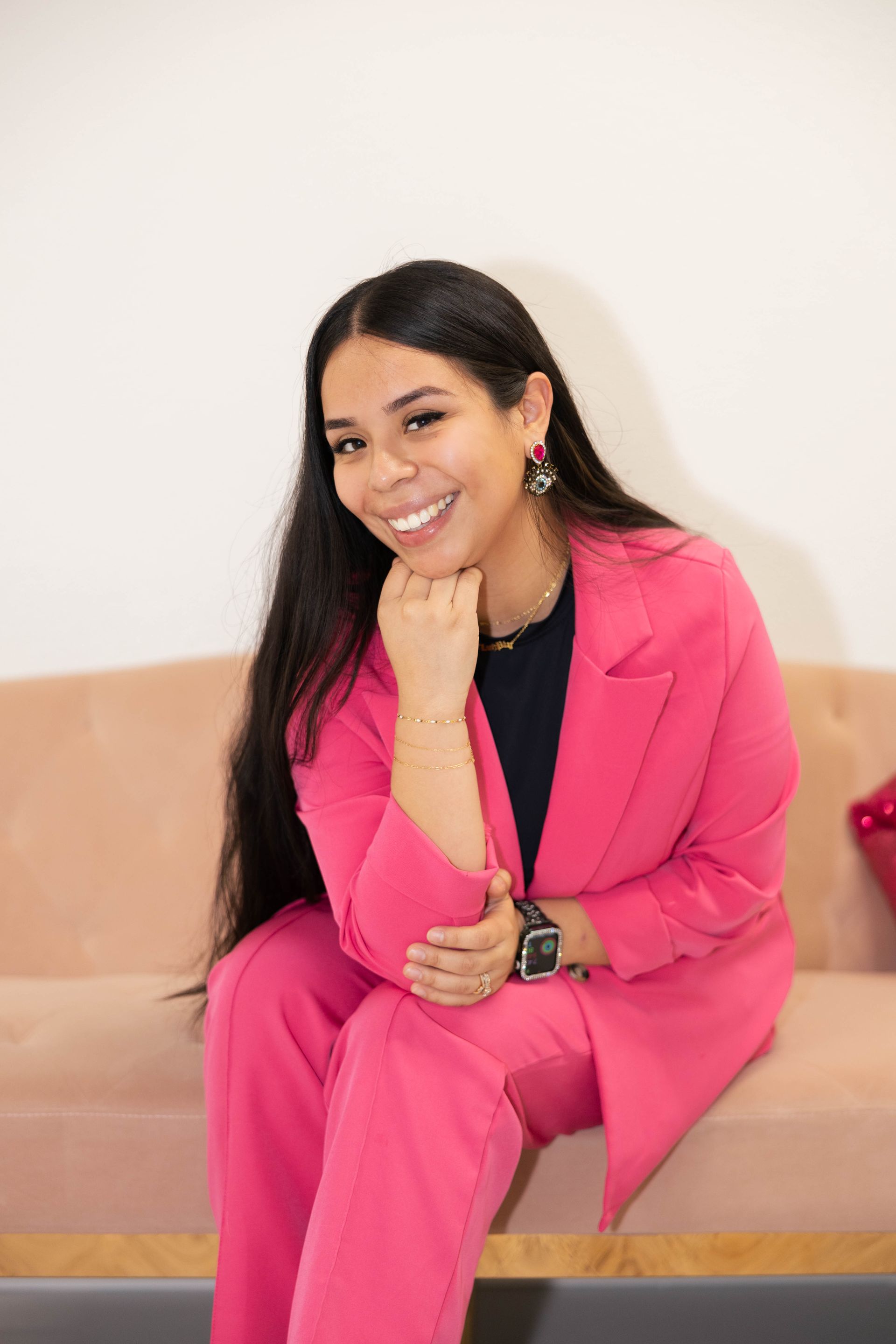 Woman in a bright pink suit seated on a beige couch, smiling with her chin resting on her hand.
